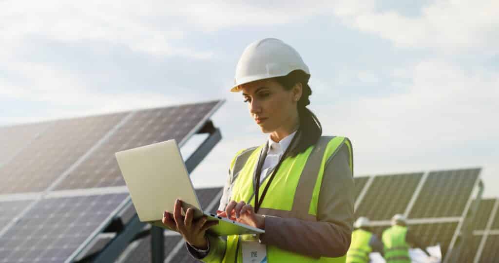 Portret inżynierki elektryczności w kasku ochronnym i mundurze, używającej laptopa do sprawdzania paneli słonecznych. Techniczka przy farmie solarnej.